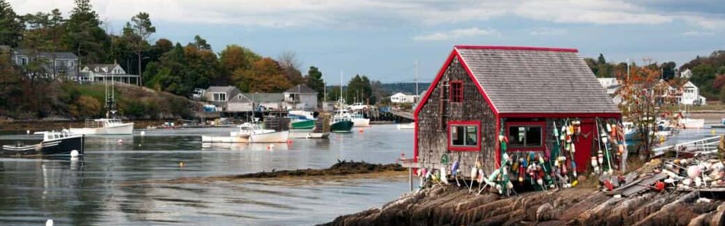 A small lobster shack covered in buoys sits at the edge of a cove filled with lobster boats.